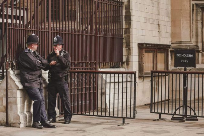 Deux policiers britanniques en uniforme sombre et casques traditionnels, portant des chaussures d'intervention noires robustes, discutant près d'une entrée réservée