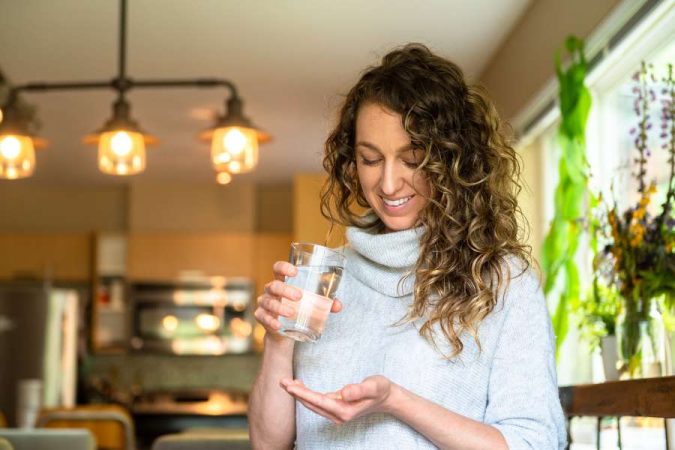 Jeune femme aux cheveux bouclés buvant un verre d'eau dans un intérieur chaleureux avec plantes vertes, illustrant les principes fondamentaux du bien-être au naturel