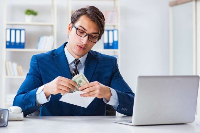 Jeune homme d'affaires en costume bleu vérifiant une enveloppe de paie contenant des billets, illustrant un aspect de la gestion de la paie en entreprise