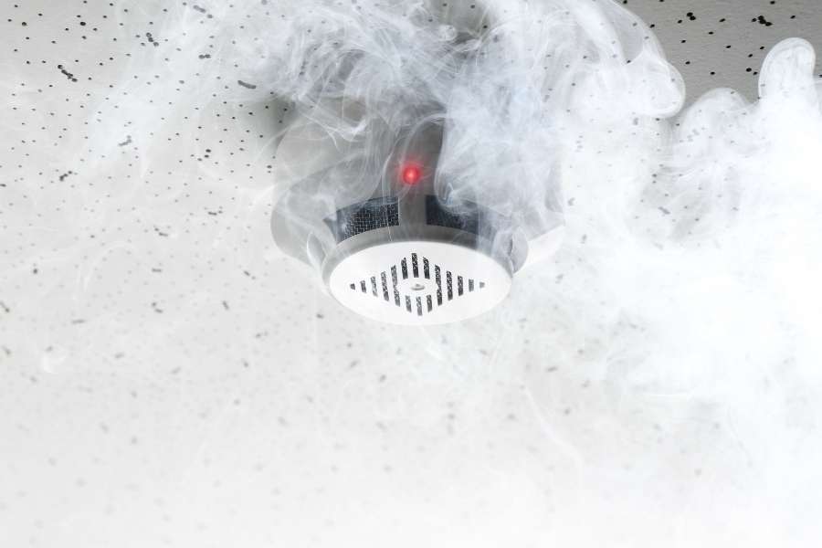 Détecteurs de fumée connectés en alerte lors d’une détection de fumée Détecteur de fumée connecté activé avec voyant rouge lumineux et fumée blanche sur plafond blanc