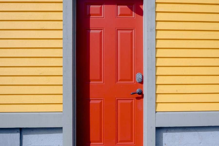 Porte d’entrée ancienne colorée rehaussant une architecture traditionnelle en bois Porte d'entrée ancienne rouge vif à panneaux sur façade jaune en bardage bois avec encadrement gris