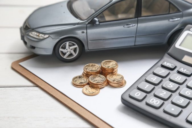 Gros plan sur une maquette de voiture grise, des pièces d'or et une calculatrice sur un bureau, évoquant le calcul pour une assurance temporaire voiture.