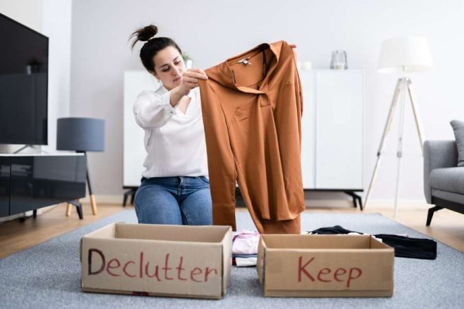 Une femme trie ses vêtements dans son salon avec deux cartons étiquetés "Declutter" et "Keep" pour désencombrer son intérieur.