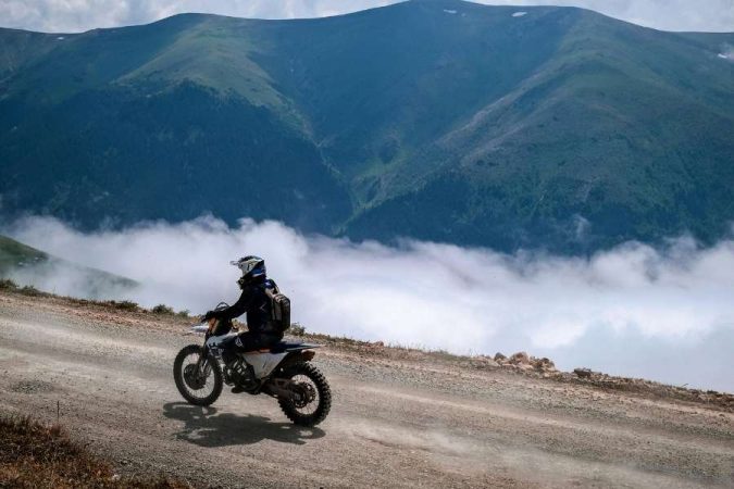 Un motard en tenue de protection pratique la conduite sur route de montagne sur un chemin de gravier, avec des nuages et des sommets verdoyants en arrière-plan.
