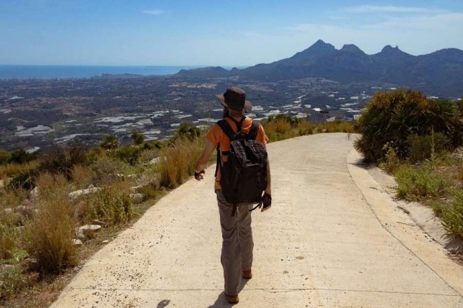 Un voyageur solitaire avec un sac à dos marche sur un sentier de montagne lors de son premier voyage en solo, avec une vue panoramique sur la mer et les montagnes.