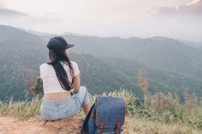 Une femme assise de dos sur un promontoire herbeux, portant une casquette et un jean, contemple un vaste paysage de montagnes brumeuses lors de son premier voyage en solitaire.