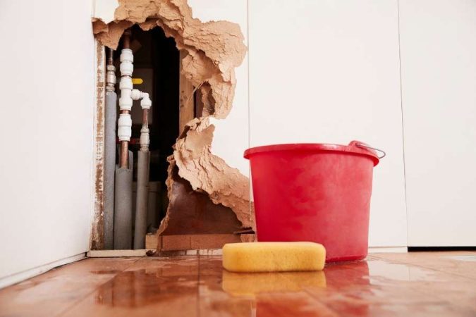 Des canalisations font un bruit de tambour suite à une fuite dans un mur endommagé avec un seau rouge.