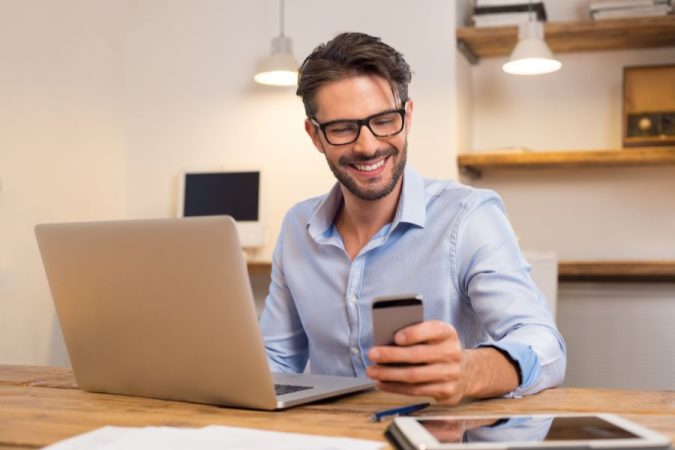 Un homme d'affaires souriant utilise son smartphone à côté de son PC portable sur un bureau en bois.